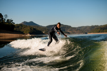 Fototapeta na wymiar flexible man in wetsuit on wakesurf masterfully riding on splashing river wave
