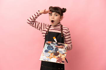 Little artist girl holding a palette isolated on pink background doing surprise gesture while looking to the side