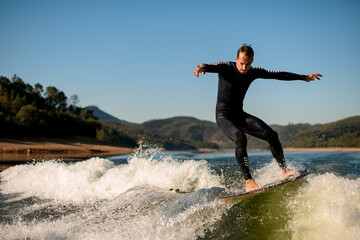 man in black wetsuit stands on splashing wave on a wakesurf