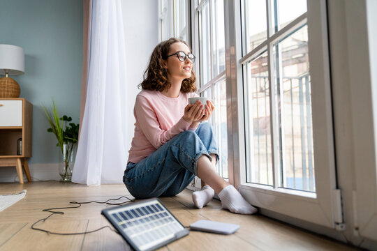 Young businesswoman with coffee cup looking through window at home