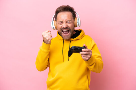 Middle age man playing with a video game controller isolated on pink background celebrating a victory in winner position