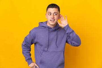 Young caucasian man isolated on yellow background listening to something by putting hand on the ear