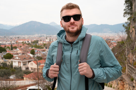 Young caucasian blond bearded middle aged man with backpack,tourist looking at camera on background of beautiful city,orange roofs of town of Fethie,Turkey, mountains.Concept of travel,tourism,trip