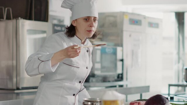 Female chef cooking gourmet soup recipe and doing taste test, making delicious vegetables meal in restaurant kitchen. Professional cook testing food sauce, feeling pleased with culinary recipe.