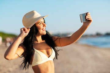 people, summer and swimwear concept - happy smiling young woman in bikini swimsuit and straw hat taking selfie with smartphone on beach