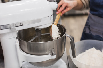 Woman adding flour with wooden spoon into mixture in blender