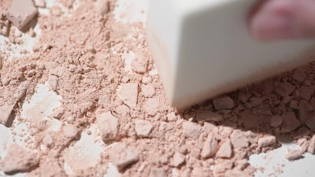Beige face powder scattered on the table. The crumbled cosmetic product is mixed by hand with a face sponge. Bright light and white background