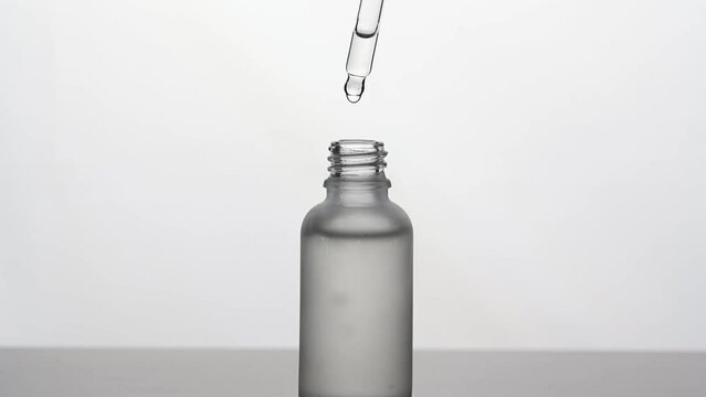 Transparent bottle with cosmetic product on white background. A drop of serum drips from a pipette into a cosmetic container