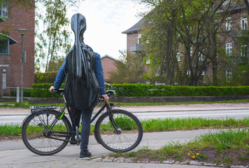 Young musician with a black cello case on his back stands with his bicycle in a suburban street in a residential area on the way to music lessons or to an apartment tour