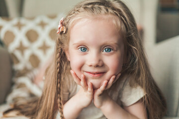 A little cute blue-eyed girl 4 years old with long hair is happy sitting in an armchair in a cozy...