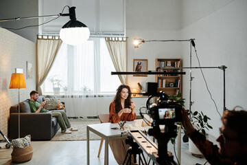 Happy young female blogger sitting by table in loft apartment during video record of commercial being shot by cameraman