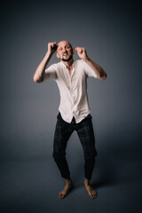 studio portrait of a bald guy with a beautiful beard barefoot in a white shirt and plaid pants dancing on a gray background