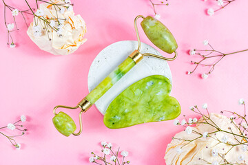 Green quartz stone gua sha scraper and jade roller for facial beauty lifting anti age massage with white flowers on pastel pink background top view.