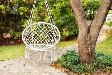 Comfortable hanging wicker white chair in summer garden. Cozy hygge place for weekend relax in garden. Hammock chair in boho style hanging on tree. Cozy exterior backyard. Concept of rest outdoor.	