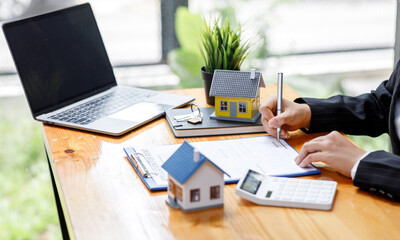 Businesswoman realtor taking notes and holding house model, sitting at desk with paper house model...