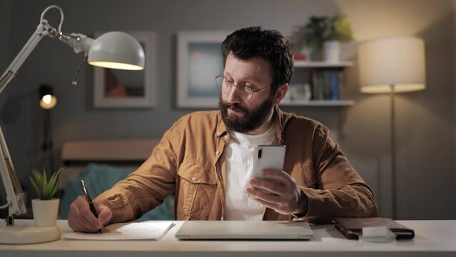 Man works using phone. Bearded freelancer in home office reading message, typing and taking notes. Remote worker manager collects and consolidates data