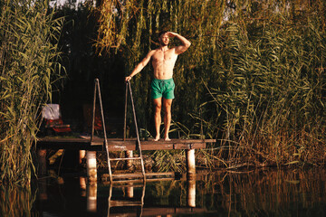 Fototapeta na wymiar Handsome shirtless young man stands on a dock on a lake and looking into the distance