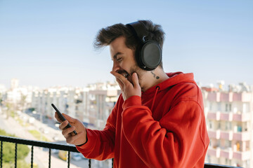 Laughing, the young man laughs loudly at the message he receives, young man wearing headphones on balcony with phone in hand, funny moment and smile. face expression