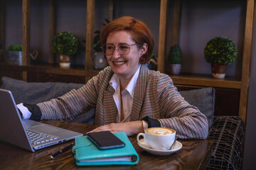 Senior red haired woman works at laptop while sitting in small cozy cafe. Diversity people.