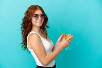 Teenager redhead girl isolated on blue background in swimsuit and holding a cocktail
