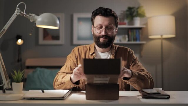 Man opens box positive emotions. Happy bearded male buyer opens received package and experiences positive emotions. Shopaholic in home office unpacking online purchase