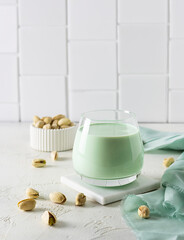 Pistachio milk in a clear glass on a marble stand. Pistachios in a white vase in the background. Light green napkin on the table. Light brick background