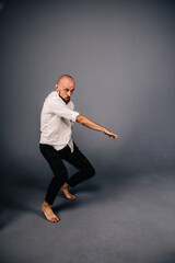 studio portrait of a bald guy with a beautiful beard barefoot in a white shirt and plaid pants dancing on a gray background