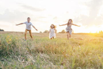 Naklejka na ściany i meble Portrait of three children playing game of catch, jumping and running on dry grass hay field paths in sunset. Trees and meadow on background. Looking around. Cloudy sunny sky. Haying time