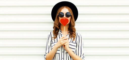 Portrait of stylish young woman with red heart shaped lollipop wearing black round hat, striped...