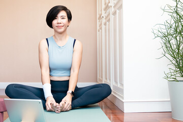 Healthy and stylish Asian woman sit in easy seat pose or sukhasana practicing yoga online class via computer tablet, look at camera and smile in bedroom at home. Wellbeing and active lifestyle.
