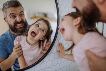 Father having fun with his little daughter, making grimaces in mirror in bathroom at home.