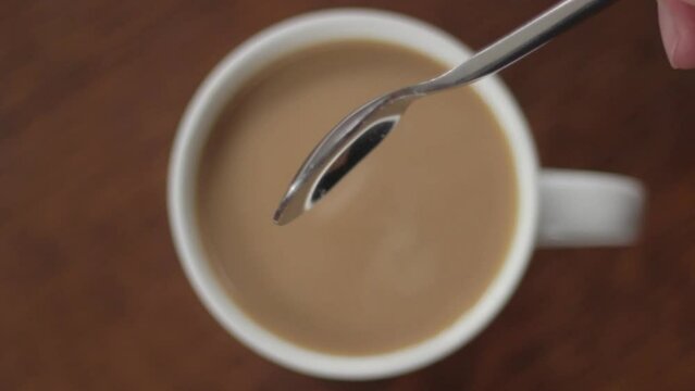 Pouring tea spoon of sugar into white coffee drink and mixing, close up top down view