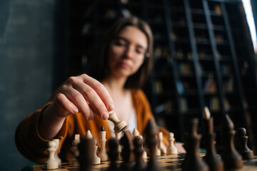 Close-up selective focus of smart young woman in elegant eyeglasses making chess move sitting in armchair in dark library room. Pretty intelligent lady playing logical board game alone at home.
