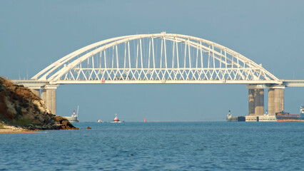 Fototapeta premium View of the beautiful large white bridge for cars moving above the calm sea and cargo ships against blue clear sky. Shot. Picturesque marine landscape