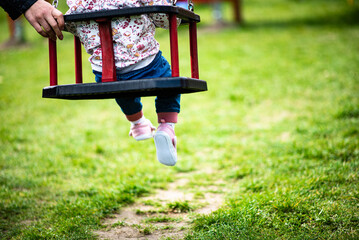 baby girl is swinging on a swing with mother hand at blurred grass background, toddler legs - obrazy, fototapety, plakaty