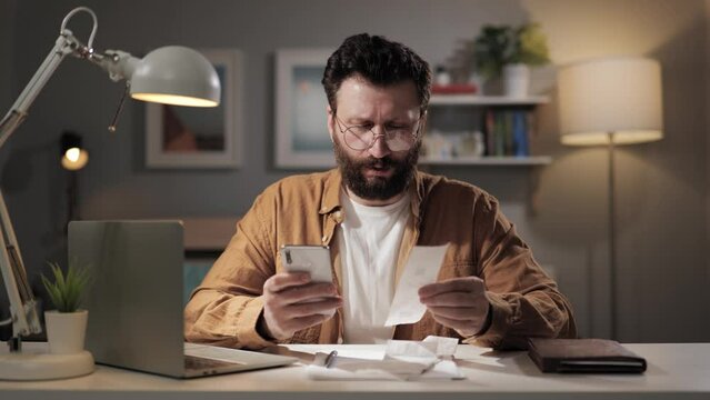 Man counts pay checks. Upset bearded man at home counts expenses on calculator in his phone. Freelancer plans to pay taxes