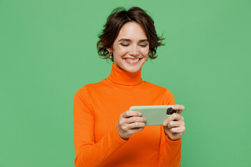 Young smiling woman 20s wear orange turtleneck using play racing app on mobile cell phone hold gadget smartphone for pc video games isolated on plain pastel light green background studio portrait.