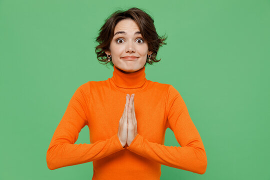 Young pleaded woman 20s in orange turtleneck hold hands folded in prayer gesture, beg about something isolated on plain pastel light green color background studio portrait People lifestyle concept