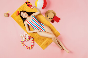 Top view full body young woman of Asian ethnicity in striped swimsuit lies on towel hotel pool do selfie shot mobile cell phone isolated on plain pastel pink background. Summer vacation sea concept.
