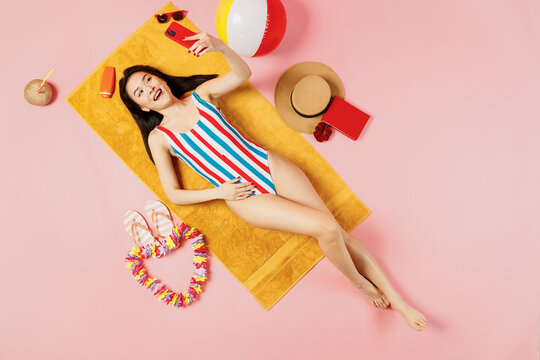 Top view full body young woman of Asian ethnicity in striped swimsuit lies on towel hotel pool do selfie shot mobile cell phone isolated on plain pastel pink background. Summer vacation sea concept. - Powered by Adobe