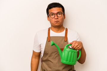 Young gardener hispanic man holding a watering can isolated on white background shrugs shoulders and open eyes confused. © Asier