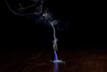 Incense cone burning with white smoke in the air and bluish smoke at the base of the cone dark...