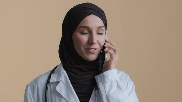 Arabian muslim young woman surgeon practitioner wear medical coat traditional hijab talk by cellphone consult patient remote female islamic therapist discuss treatment mobile conversation at clinic