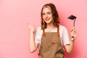 Young gardener woman holding gardening shovel isolated on pink background showing a copy space on a palm and holding another hand on waist.