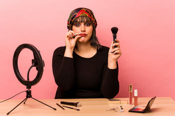 Young makeup artist woman doing a makeup tutorial isolated on pink background with fingers on lips keeping a secret.