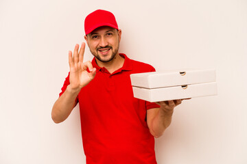 Young delivery man holding pizzas isolated on beige background cheerful and confident showing ok gesture.