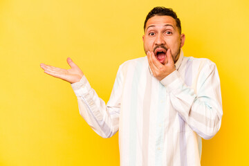 Young hispanic man isolated on yellow background holds copy space on a palm, keep hand over cheek. Amazed and delighted.