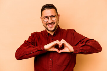 Young hispanic man isolated on beige background smiling and showing a heart shape with hands.