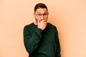 Young hispanic man isolated on beige background covering mouth with hands looking worried.