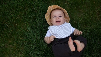 Small Happy Newborn child in summer panama hat Fall down laying on grass barefoot in Summer Sunny Day. Infant Kid Toddler Boy Smilling Face look at camera in Garden ouside Family Childhood Nature.